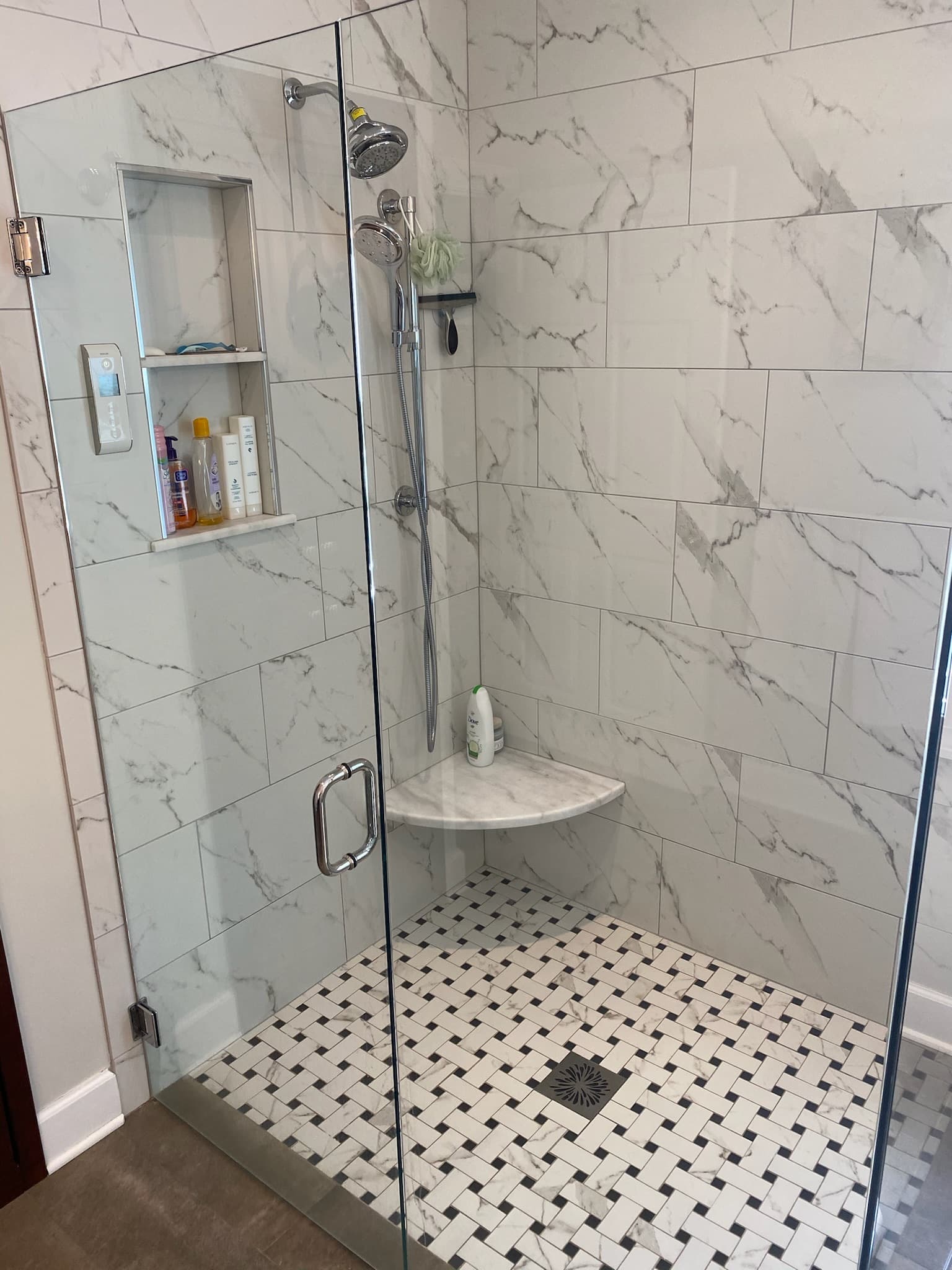 A walk-in shower with white marble-look subway tile walls, a patterned black and white tile floor, and chrome fixtures