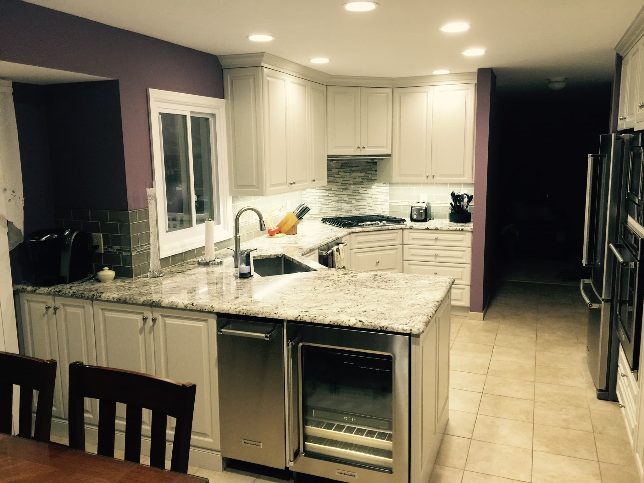 Wide view of a transitional kitchen with white recessed panel cabinets, granite countertops, and a central island with a wine fridge