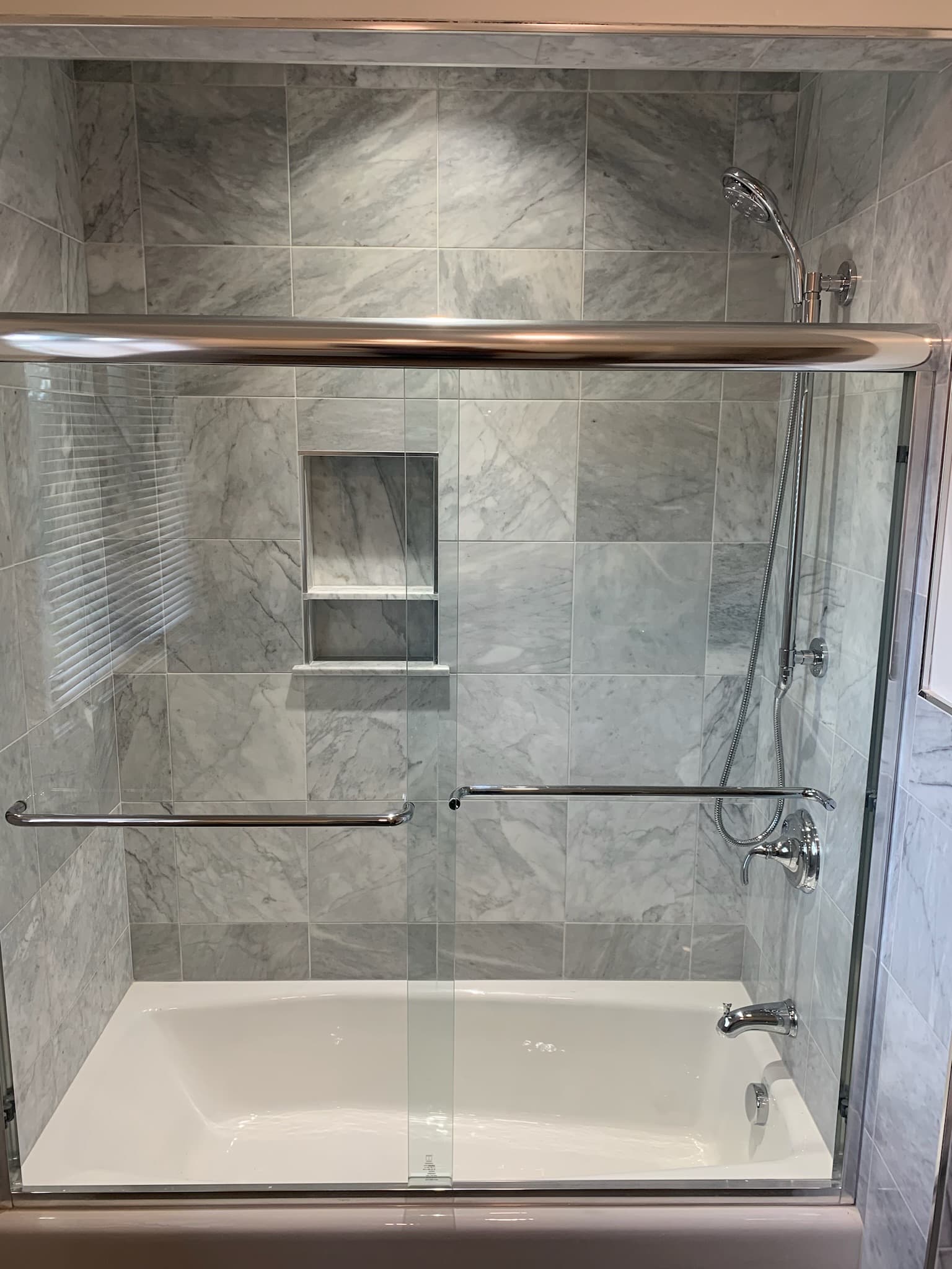 A bright transitional bathroom features a tub-shower combo with gray marble-look tile walls and a chrome sliding glass door