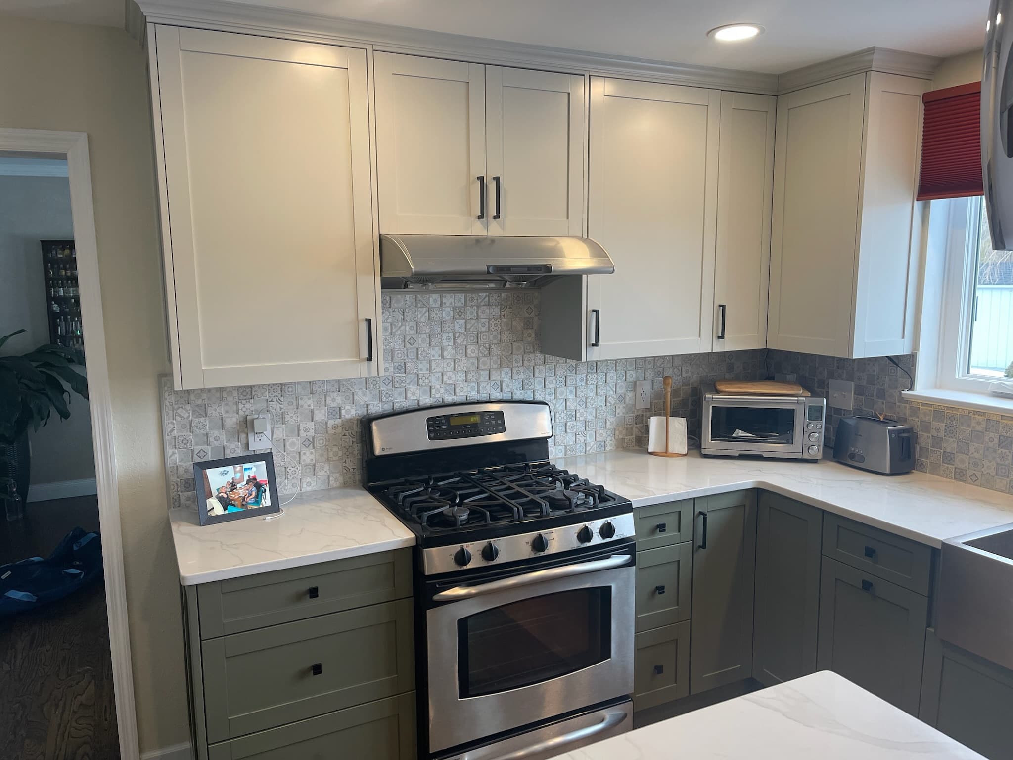 A transitional kitchen features two-tone shaker cabinets in green and white, a stainless steel range and hood, and patterned tile backsplash