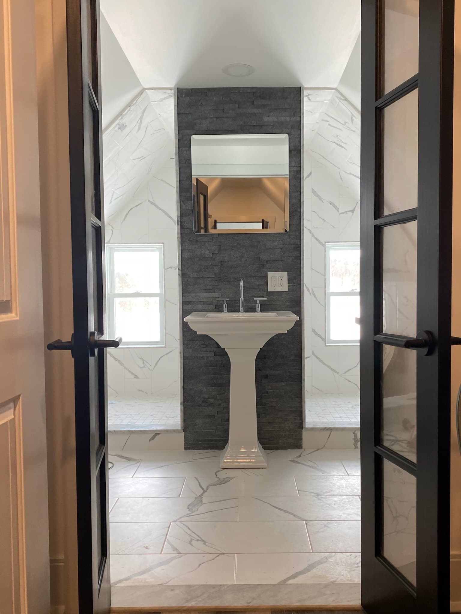 A contemporary bathroom with a white pedestal sink in front of a dark gray stone accent wall and white marble-patterned tiles