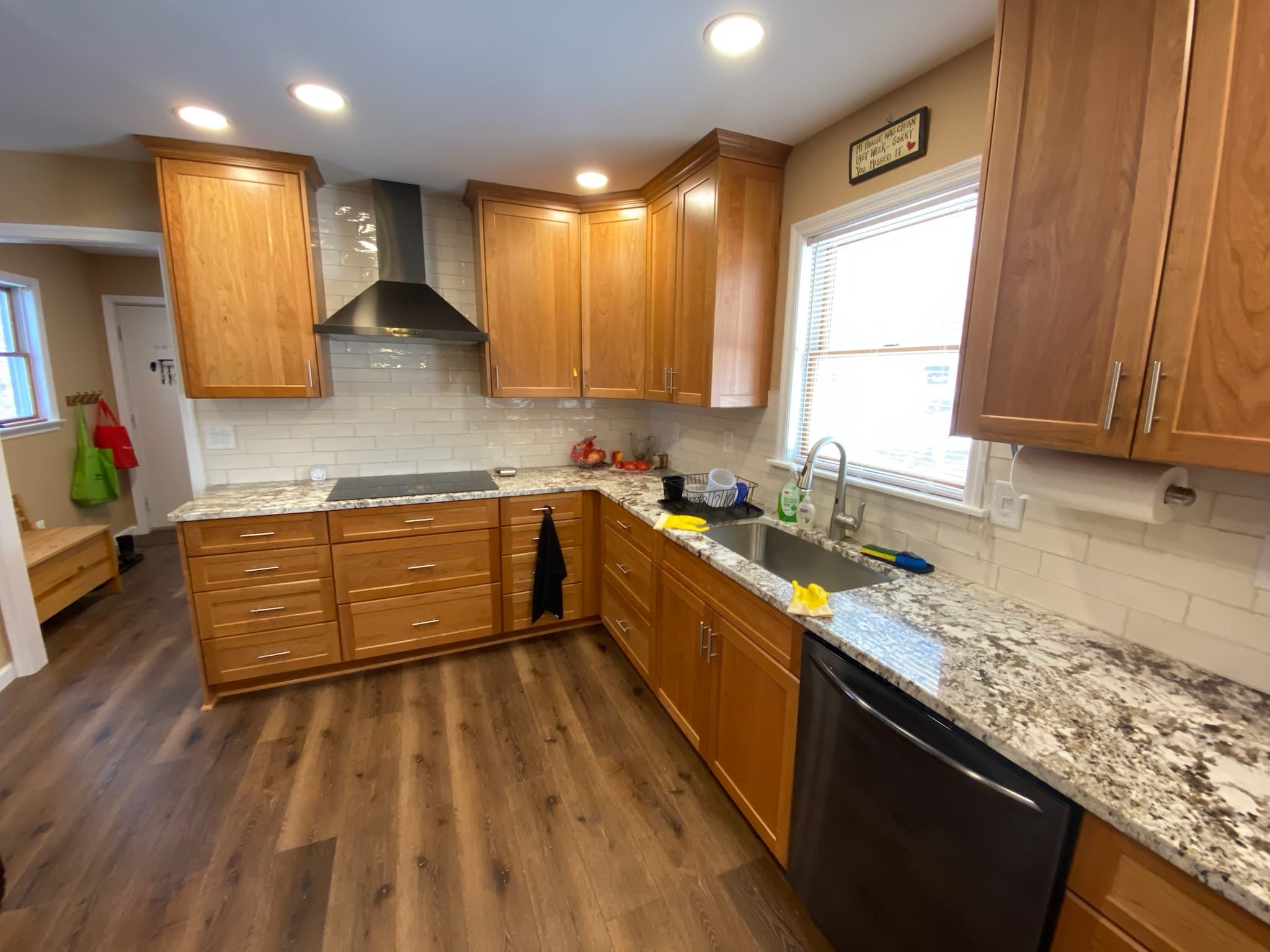 Wide view of a transitional kitchen with medium wood flat panel cabinets, granite countertops, and a black range hood over a cooktop