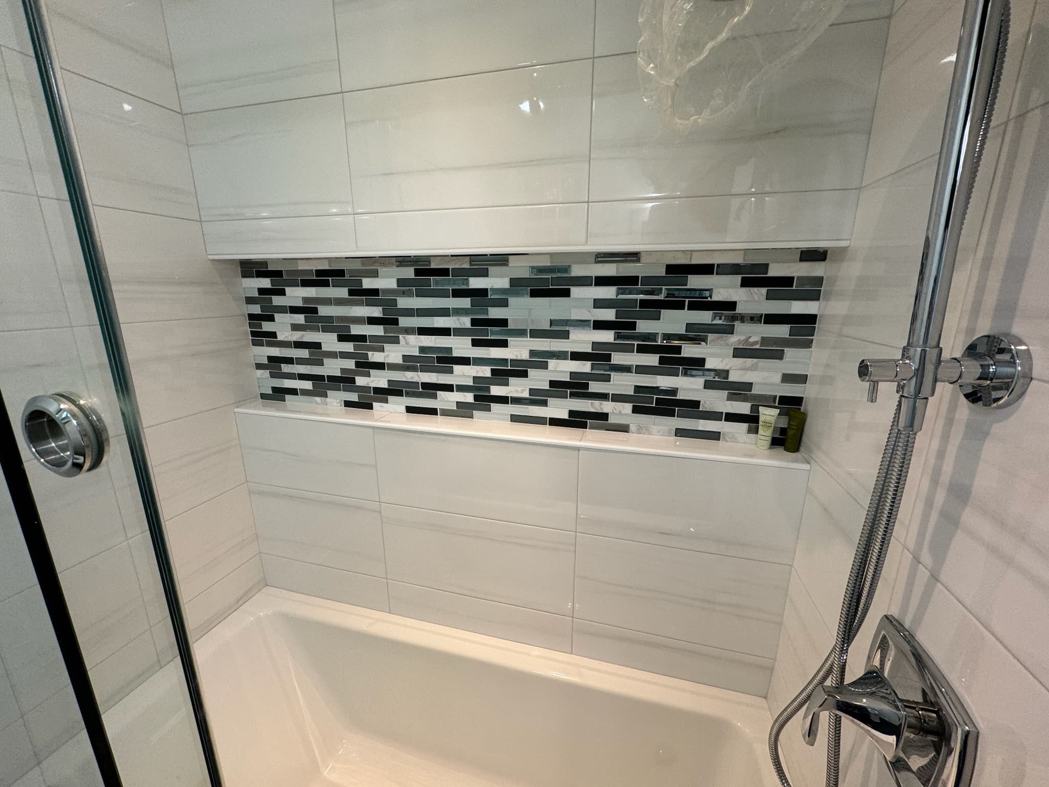 A bright contemporary bathroom features a white tub-shower combo with a recessed shelf accented by black, white, and gray mosaic tiles