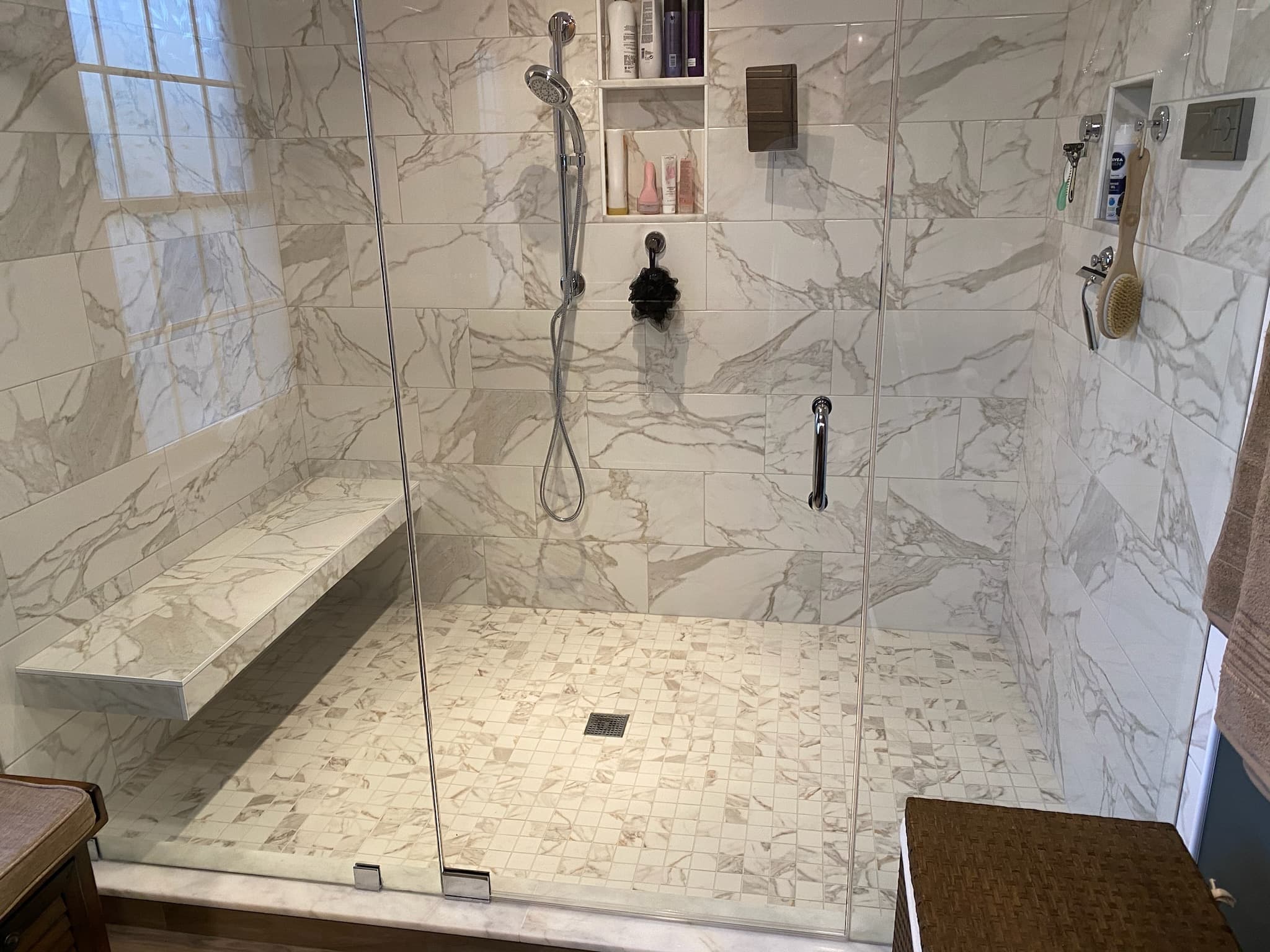 A bright, spacious shower features large format marble-look tiles, a mosaic tile floor, and a built-in bench