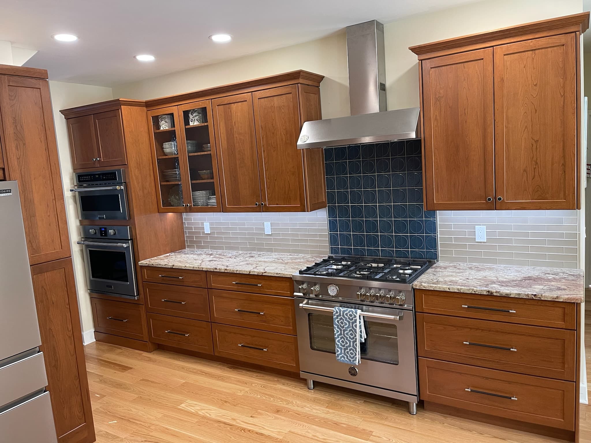 Transitional kitchen with medium wood recessed panel cabinets, granite countertops, and a modern range hood above a patterned blue tile backsplash